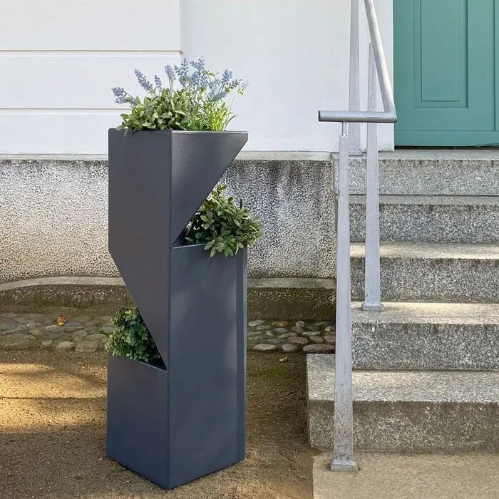 Ein moderne Blumensaeule mit geometrischem Design, üppigen grünen Pflanzen und violetten Blumen, neben einer Treppe und einer blaugrünen Tür platziert.