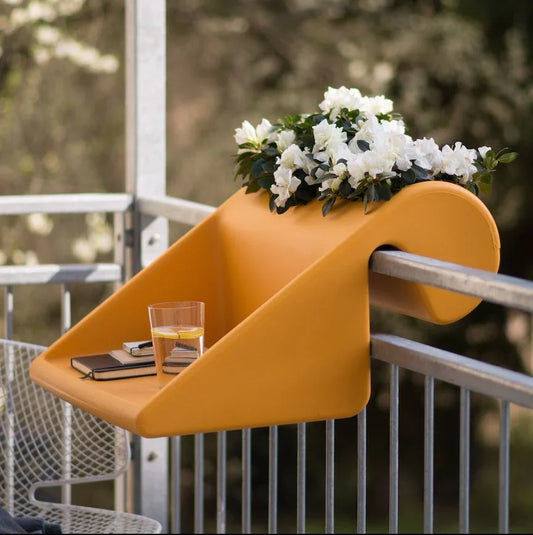 Auf einem leuchtend orangefarbenen Balkontisch stehen ein Glas Wasser, ein Notizbuch und ein Topf mit weißen Blumen und verschönern den Außenbereich.