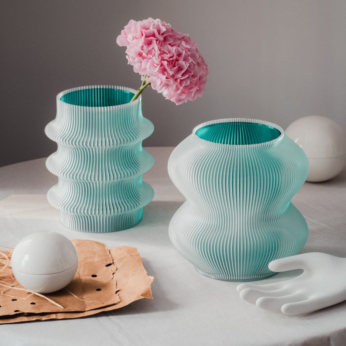 Zwei stilvolle Vasen mit strukturiertem mintgrünem Muster halten eine rosa Blume. Eine weiße Kugel und eine Handskulptur stehen in der Nähe auf einem neutralen Tisch.