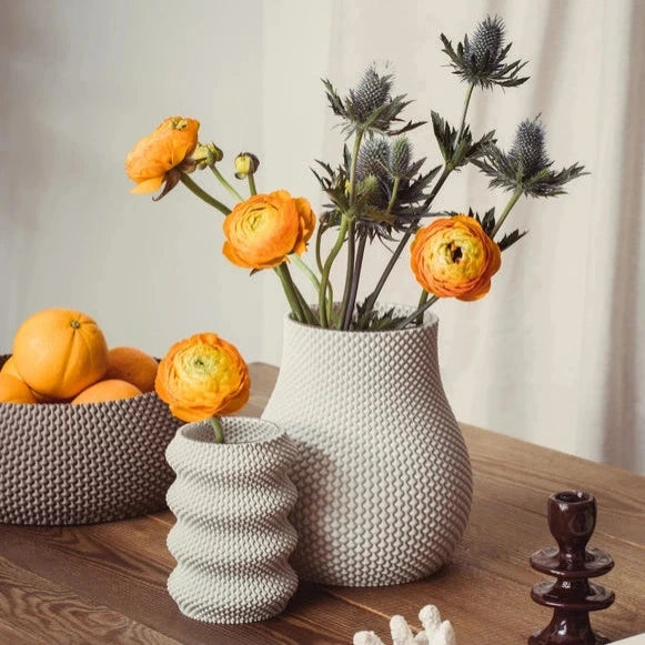 Auf einem Holztisch stehen strukturierte Vasen mit leuchtend orangefarbenen Ranunkeln und Disteln, daneben Schalen mit Orangen und dekorative Akzente.