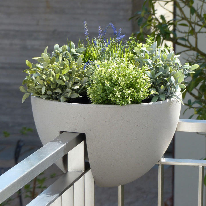 Ein moderner, runder Blumentopf, gefüllt mit üppigen grünen Kräutern und kleinen violetten Blumen, aufgehängt an einem Balkongeländer.